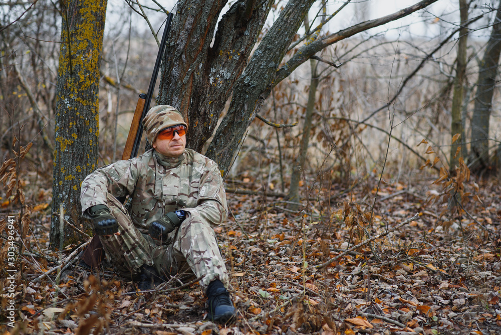 Obraz premium A male hunter with a gun while sitting takes aim at a forest. The concept of a successful hunt, an experienced hunter. Hunting the autumn season. The hunter has a rifle and a hunting uniform.