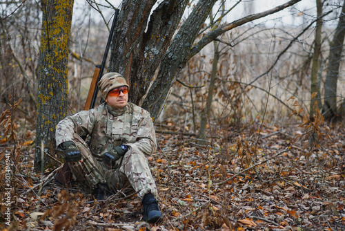 A male hunter with a gun while sitting takes aim at a forest. The concept of a successful hunt, an experienced hunter. Hunting the autumn season. The hunter has a rifle and a hunting uniform.