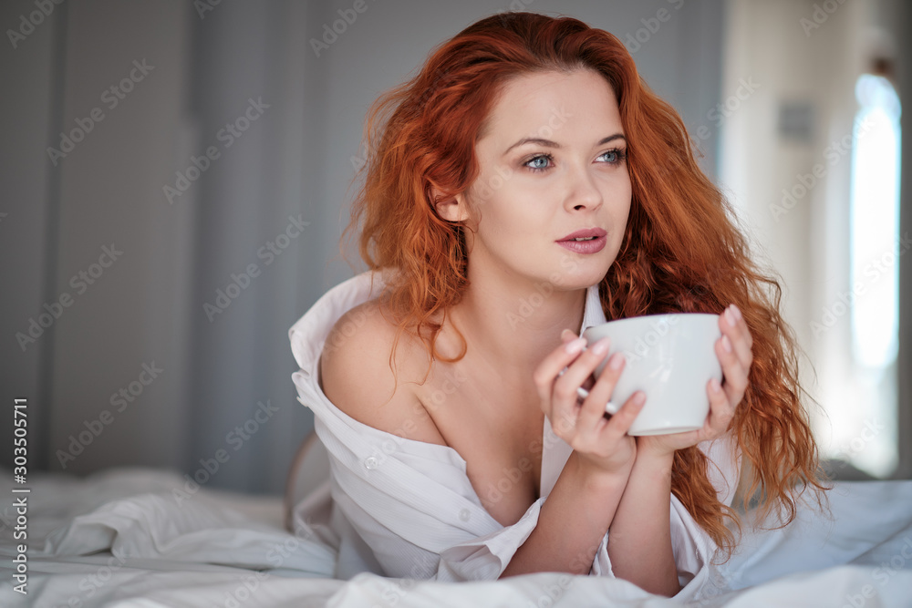 Attractive redhaired woman with a cup of coffee on the bed