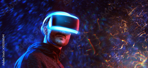 Young man on virtual reality background. Guy using VR helmet. Augmented reality, future technology, game concept. Blue neon light, fire flares.