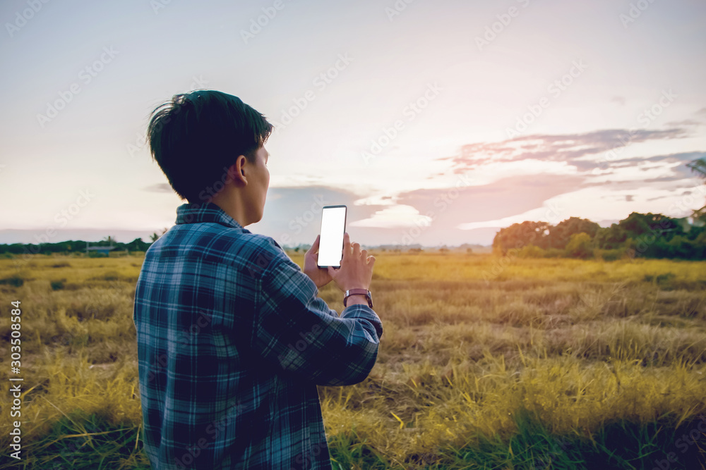 Φωτογραφία Half Back view, Smiling Asian man wearing plaid shirts and vintage eyeglasses are using a smartphone, Screen white space mock-up in evening with sunset outdoor