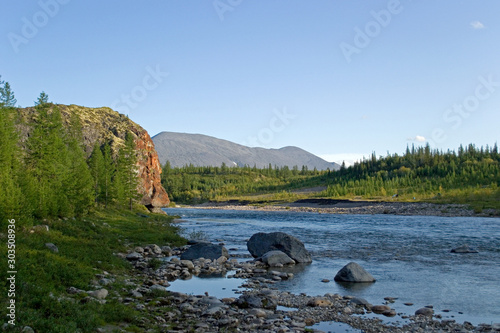 Kozhim River. A high cliff on one side of the river and a gentle wooded shore on the other side. A mountain is visible in the distance. A clear sunny day. Subpolar Ural, Yugyd Va National Park photo