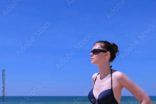 Portrait young woman sunbathing stand on beach in swimsuit on sea and blue sky background. Brunette girl is in sunglasses and bikini, side view. Travel tourist summer season vacation.