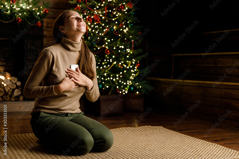 A young girl holds a mobile phone, writes a message, uses the Internet on the background of a Christmas tree and a fireplace. A young girl holds a mobile phone, writes a message, uses the Internet on the background of a Christmas tree and a fireplace.