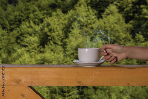 Steam rises over a cup with a hot drink in a female hand on a wooden deck against the background of the forest on a summer sunny day. Camping concept.