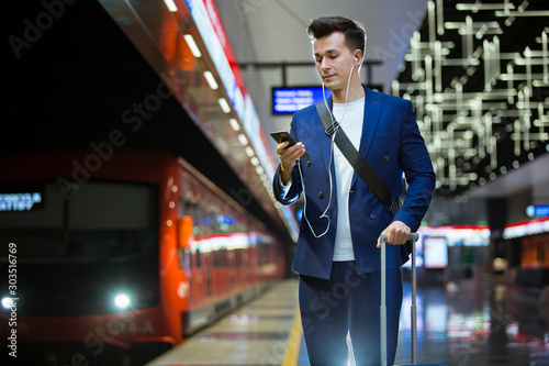 Wallpaper Mural Young stylish handsome man in suit with suitcase standing on metro station holding smart phone in hand, scrolling and texting, smiling and laughing. Futuristic bright subway station. Finland Torontodigital.ca