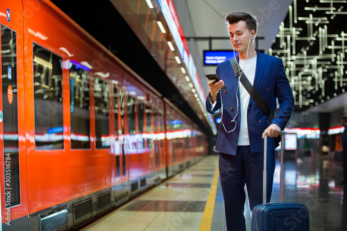 Young stylish handsome man in suit with suitcase standing on metro station holding smart phone in hand, scrolling and texting, smiling and laughing. Futuristic bright subway station. Finland photo