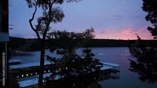 Sunrise over Lake of the Ozarks, Missouri. Two boats drive through, making wide curves in the lake. There is a covered boat dock with lights reflecting on the water. Clouds blow slowly as the sun glow