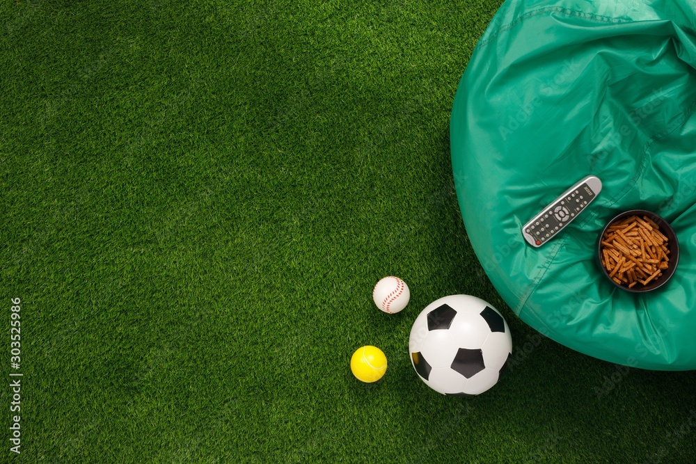 Naklejka premium Soccer ball on a green field and ottoman for a fan with snacks and a TV remote control. The apartment was lying. The concept of football matches.