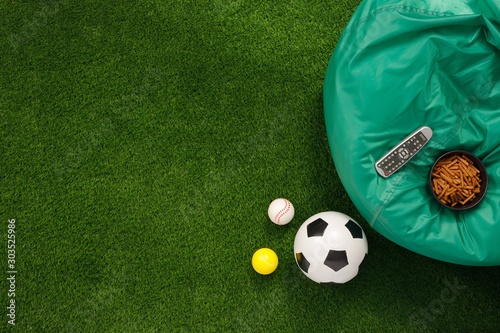 Soccer ball on a green field and ottoman for a fan with snacks and a TV remote control. The apartment was lying. The concept of football matches.