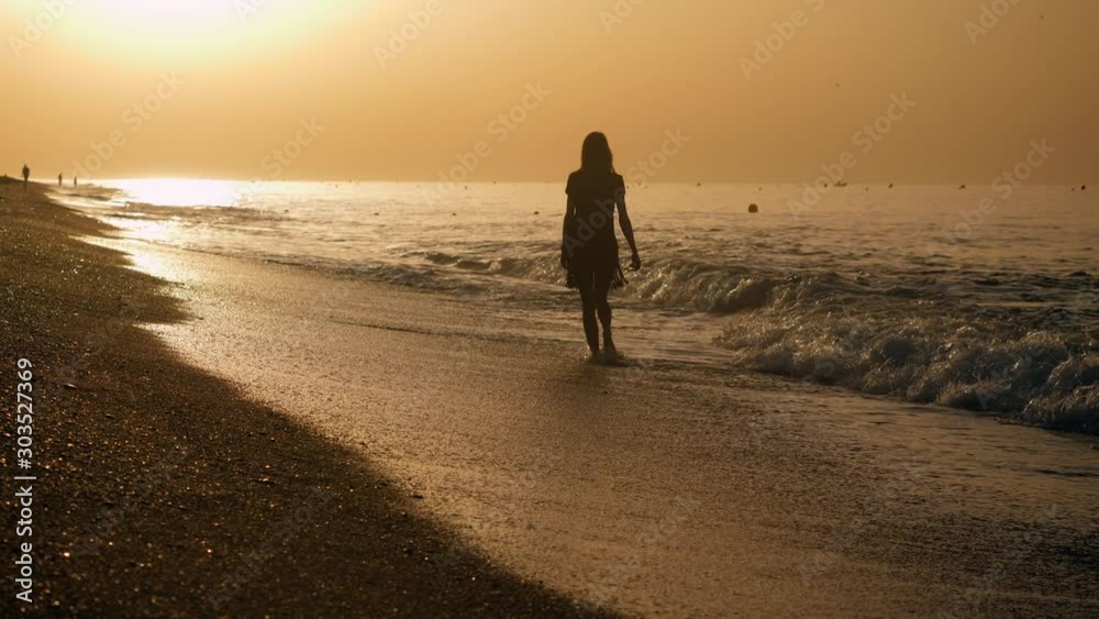 Coastline Costa del Maresme. Young beautiful woman walking barefoot across the sandy beach of Mediterranean sea at sunrise. Spain. 4K Coastline Costa del Maresme. Young beautiful woman walking barefoot across the sandy beach of Mediterranean sea at sunrise. Spain. 4K