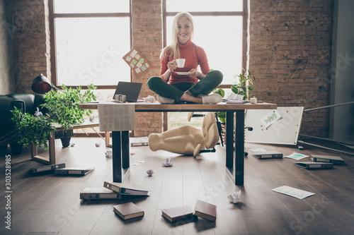 Full body photo of happy trader woman sit on table crossed legs feel carefree careless rest relax hold white coffee cup in messy office loft photo