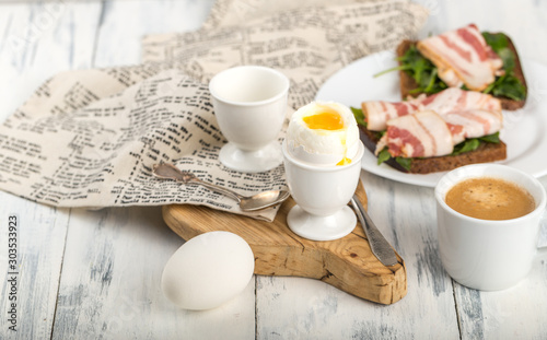 Two boiled eggs, in a shell, a drop of yolk, in a white egg stand, on a wooden Board, a textile napkin, toast with bacon on a plate, a Cup of coffee, light gray background