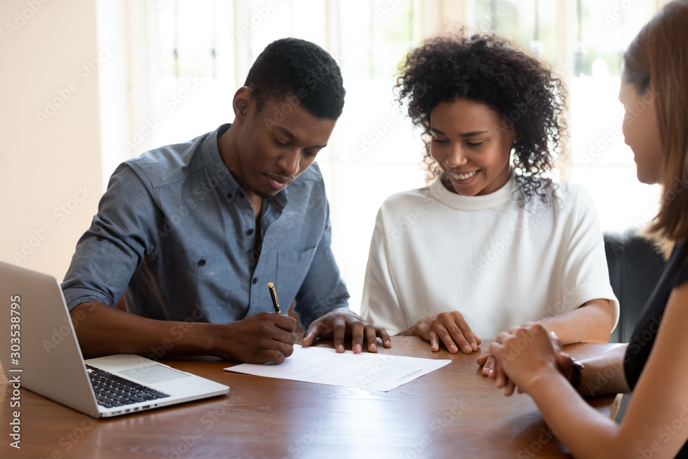 Happy young banking worker watching african ethnicity client signing agreement. Happy young banking worker watching african ethnicity client signing agreement.