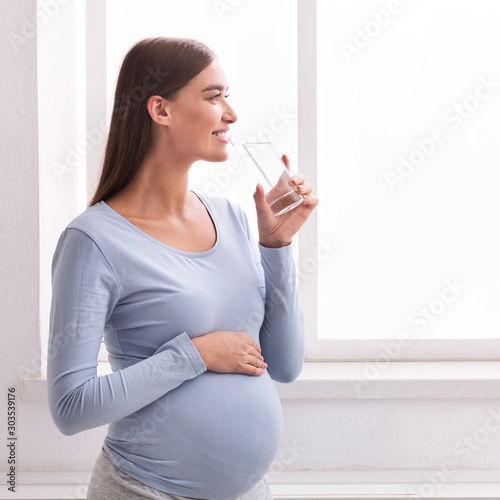 Positive Pregnant Girl Drinking Water Standing Looking In Window Indoor photo