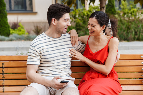 Image of happy couple laughing and using cellphone while sitting