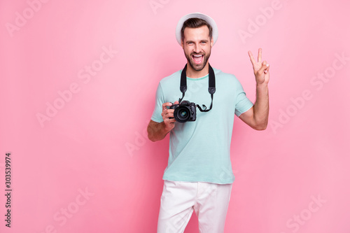 Portrait of funky funny man tourism lover have spring vacation hold dlsr professional camera make v-sign wear blue t-shirt white pants isolated over pink color background photo