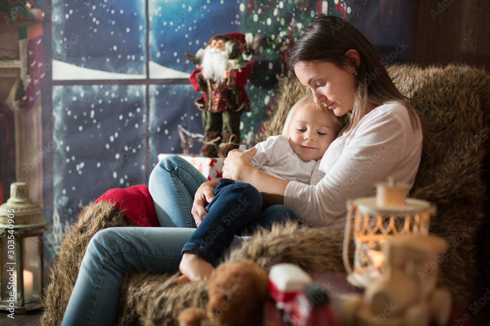 Beautiful young mother, hugging her toddler boy, sitting in cozy chair on Christmas - obrazy, fototapety, plakaty 