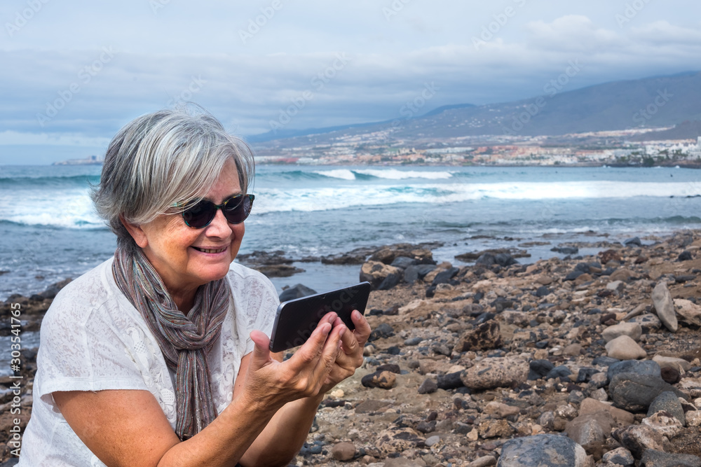 An elderly attractive lady sitting on the beach and looking at cellphone. Sea and mountain in background. Cloudy sky