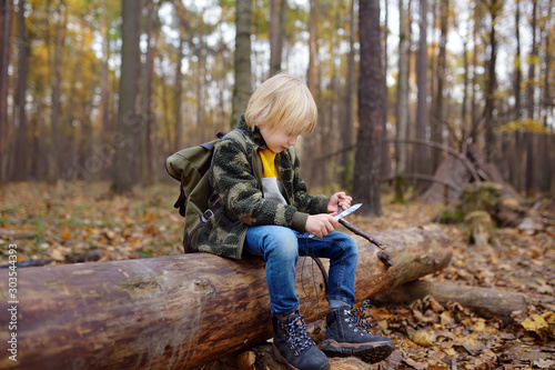 Little boy scout is sharpening a stick with the help knife in the forest.
