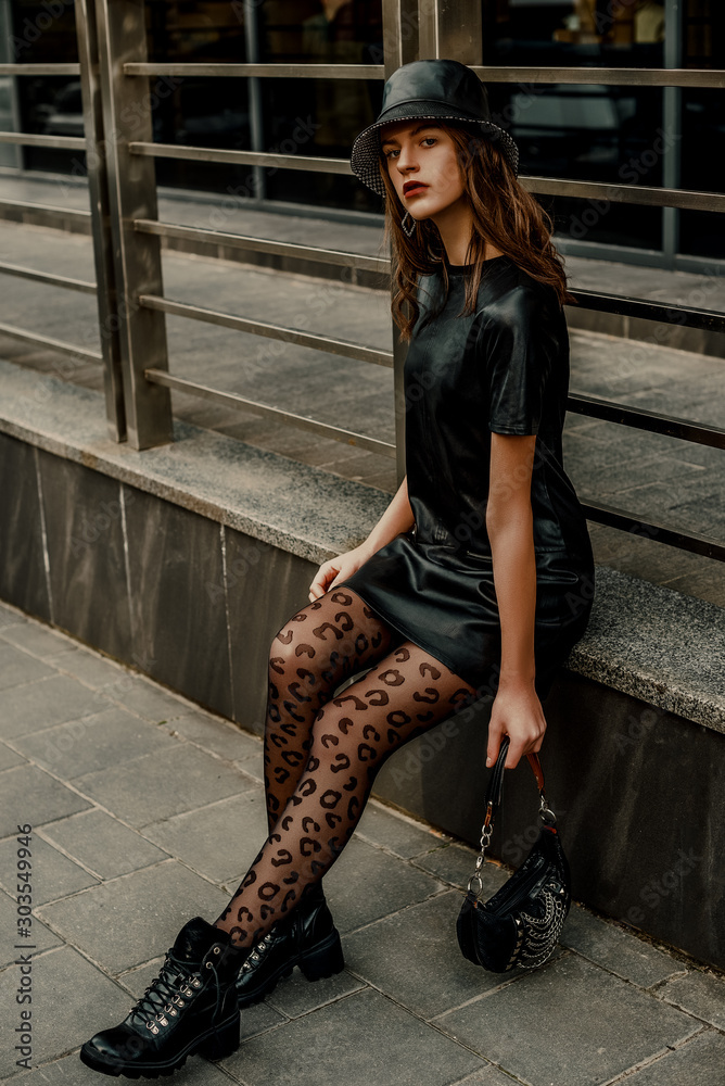 Outdoor full body street fashion portrait of young elegant model, woman  wearing trendy leather bucket hat, short dress, black leopard print tights,  lace up ankle boots, holding small stylish handbag Stock Photo