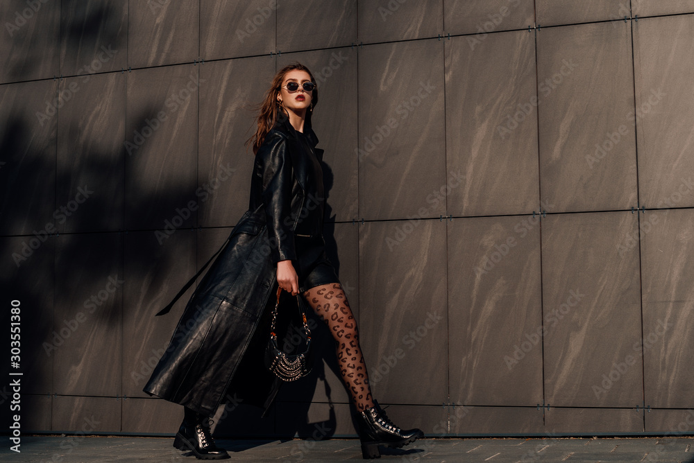 Outdoor full-length fashion portrait of young confident woman wearing total  black leather outfit, leopard print tights, holding small bag, walking in  city street, grey urban background. Copy space Stock Photo