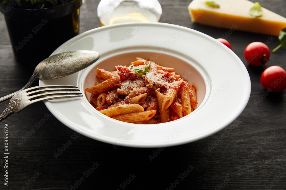 selective focus of tasty bolognese pasta with tomato sauce and Parmesan in white plate near ingredients and cutlery on black wooden background selective focus of tasty bolognese pasta with tomato sauce and Parmesan in white plate near ingredients and cutlery on black wooden background