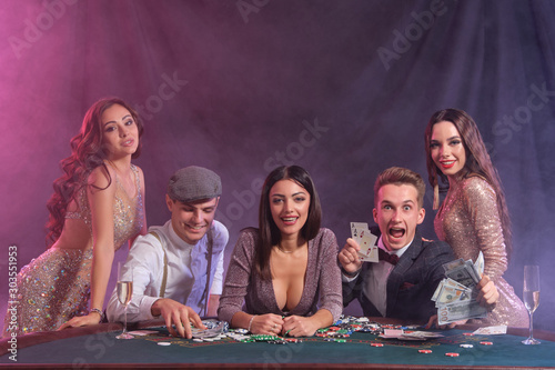 Friends playing poker at casino, at table with stacks of chips, money, cards on it. Celebrating win, smiling. Black, smoke background. Close-up.