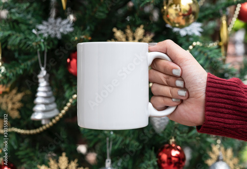 women hand holding white ceramic coffee cup on christmas tree background. mockup for creative advertising text message or promotional content. photo