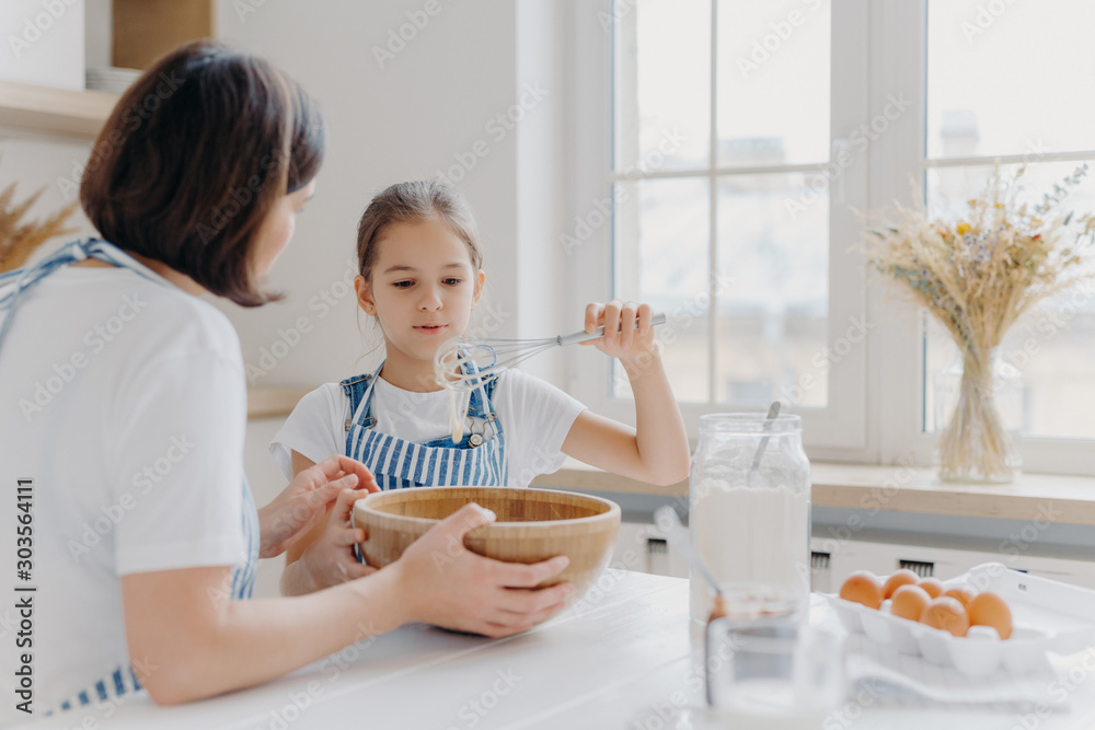 Adorable little child in apron shows whisk with white cream, wears apron, cooks together with mommy, pose in spacious kitchen together, make cake for special event. Children, help about house