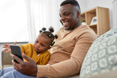 family, fatherhood and technology concept - african american father with smartphone and little baby daughter playing with ball at home