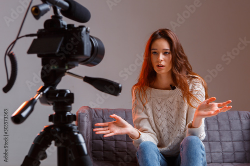 Young teen girl vlogger makes a video stream on the couch at home. A video camera on a tripod. © Georgii