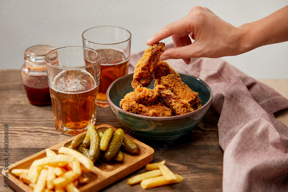 cropped view of woman eating delicious chicken nuggets, french fries and gherkins near glasses of beer on wooden table isolated on grey - obrazy, fototapety, plakaty 