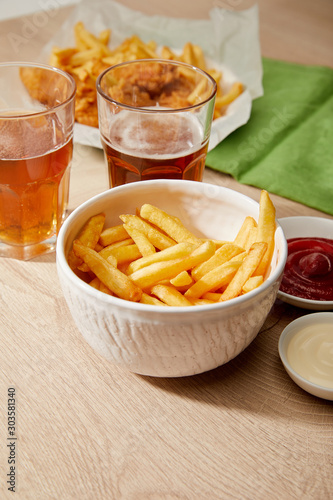 glasses of beer, french fries in bowl, sauces on wooden table