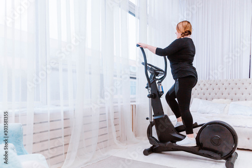 Model - a fat woman, trying to lose weight and at home is engaged in an elliptical trainer photo