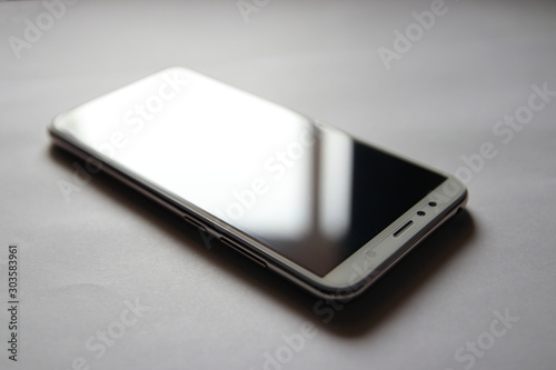 A smartphone with a protective film on the screen reflects sunlight. The modern business phone lies against a grey background.