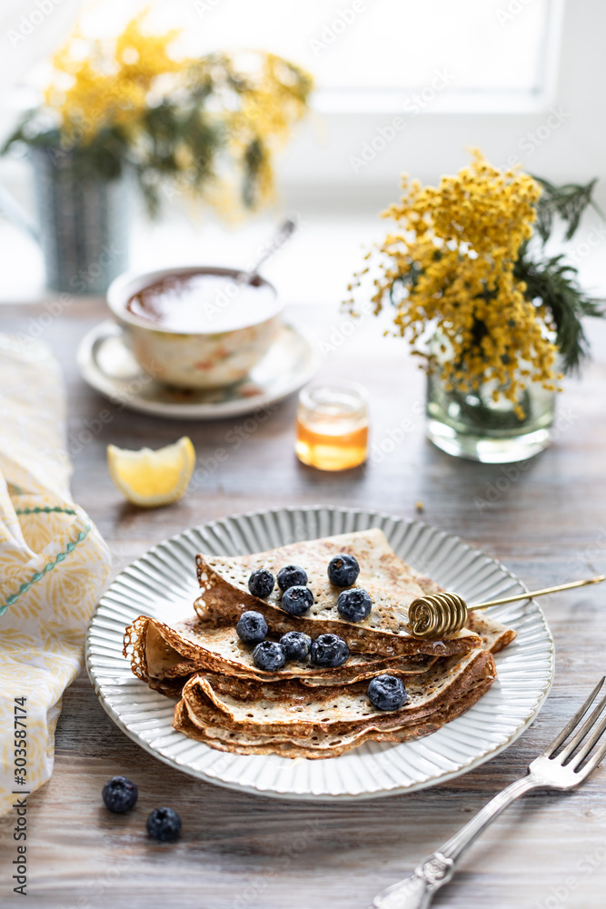 Fototapeta premium A plate with pancakes with blueberry berries on a wooden table. In the background is a cup of tea and a bouquet of spring flowers by the window.