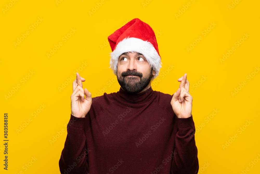 Man with christmas hat over isolated yellow background with fingers crossing and wishing the best Man with christmas hat over isolated yellow background with fingers crossing and wishing the best