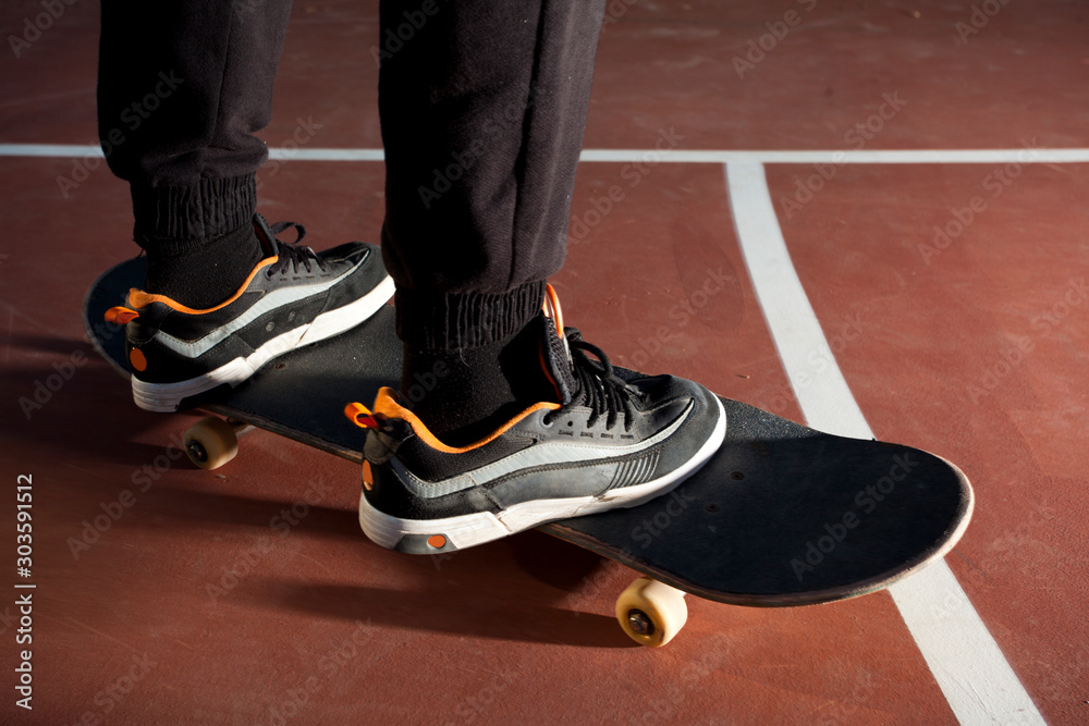 Young individual legs of the male skateboarder standing on skateboard in an urban area in a skatepark with long black pants and sneakers