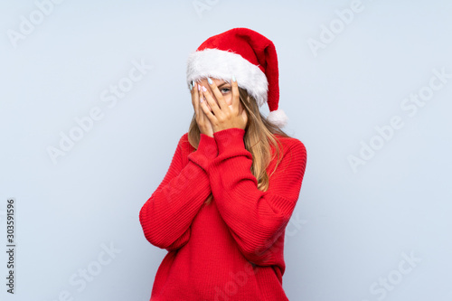 Girl with christmas hat over isolated blue background covering eyes and looking through fingers