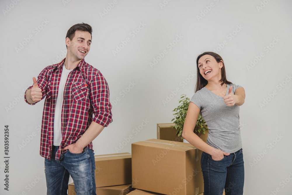 Happy man and woman standing near stack of carton boxes in their new apartment, giving thumbs up, putting one hand into pocket, smiling. Happy man and woman standing near stack of carton boxes in their new apartment, giving thumbs up, putting one hand into pocket, smiling.