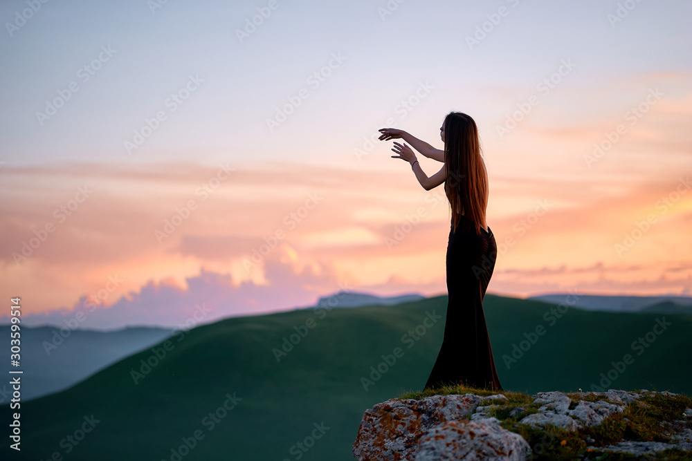 Φωτογραφία A beautiful young girl with a bare back stands in an evening dress in the mountains of the Caucasus, admiring the sunset