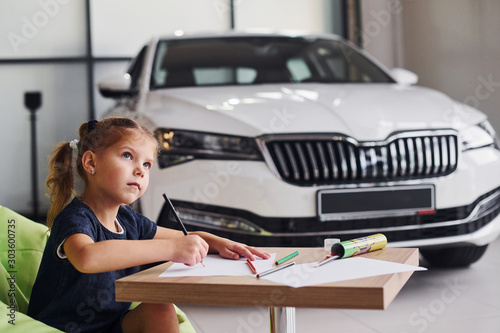 Cute little girl sits on the soft green chair by the table with pencil and paper sheets. Near modern automobile