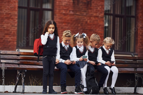 Group of kids in school uniform sits on the bench outdoors together near education building