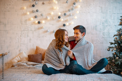 Young man looking to in to his girlfriend eyes smiling sitting with crossed legs on a bed with decorated tree and window on background. Happy loving couple enjoying Christmas. photo