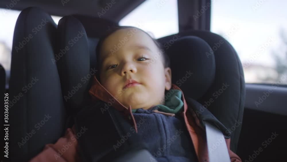 Medium shot of adorable toddler boy sitting in car seat of moving car and looking at camera