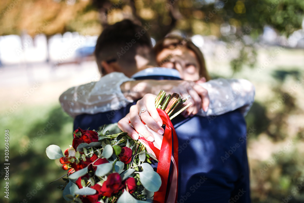 Fototapeta premium Engagement ring on the bride's finger. The bride and groom gently hug each other. Beautiful bride in a white dress with an elegant wedding ring on her finger holds a wedding bouquet of flowers. 