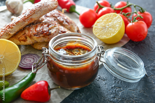 Jar of tasty barbecue sauce, chicken, vegetables and spices on table