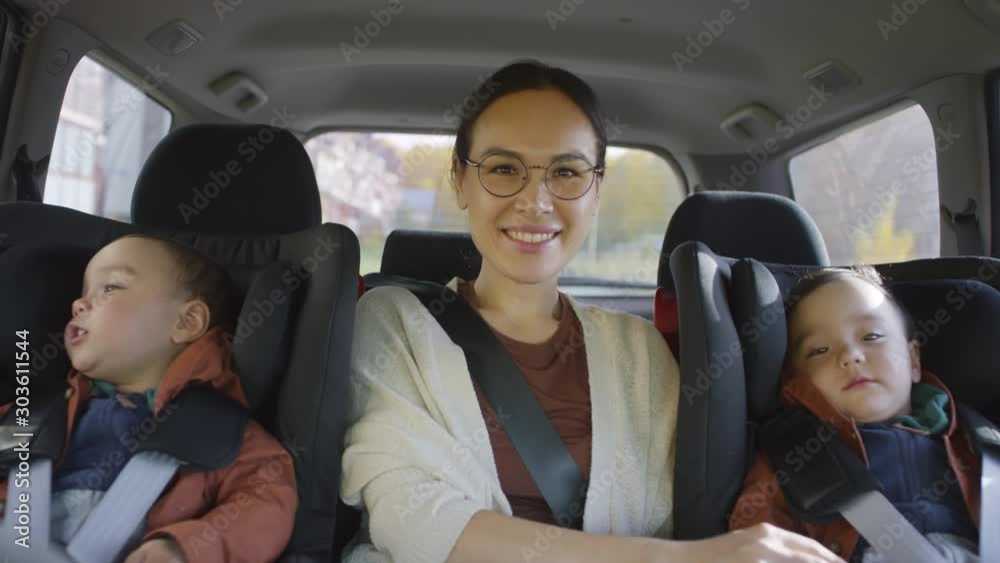 Medium shot of happy young Asian mother in glasses sitting on passenger seat of moving car with toddler boys and smiling for camera