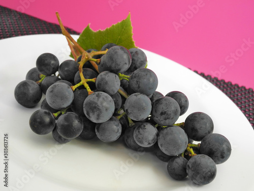 Dark purple Isabella grape bunch with leaf on white plate  on pink blackbackground photo